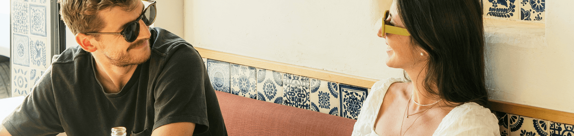 Man and woman smiling at each other in a sunlit café with patterned tile wall